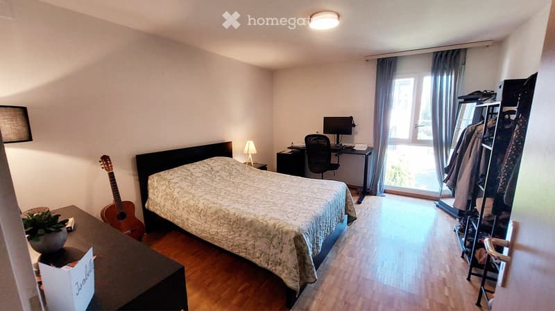 Cozy bedroom with wooden flooring, a double bed with patterned bedding, a desk with a computer, a guitar, a small plant, and a wardrobe with hanging clothes.
