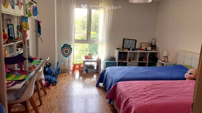 Brightly lit children's bedroom with two single beds, a desk, shelves, and various toys and decorations.