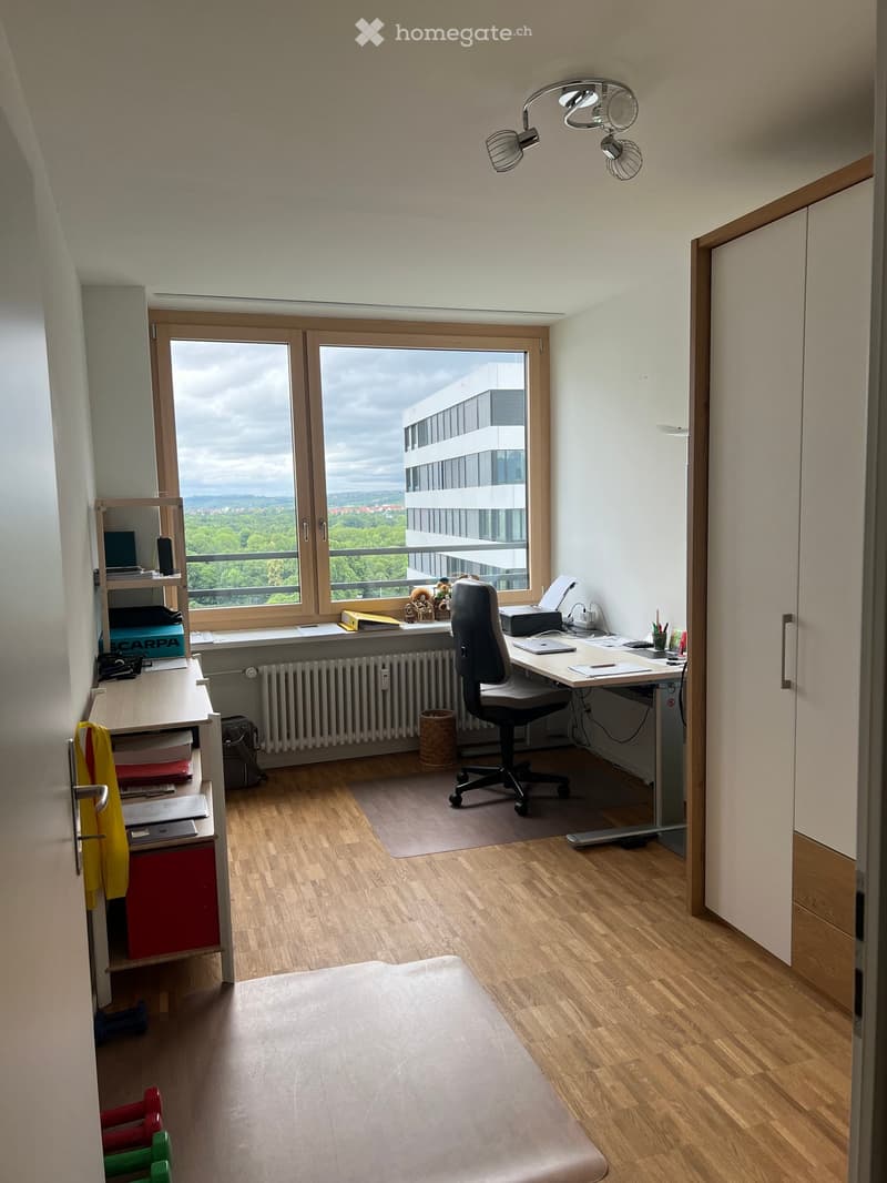 A well-lit room with wooden flooring, a large window offering a view, a desk with a chair, a laptop, printer, and various office supplies, a shelf with books and a toy, and a radiator below the window.