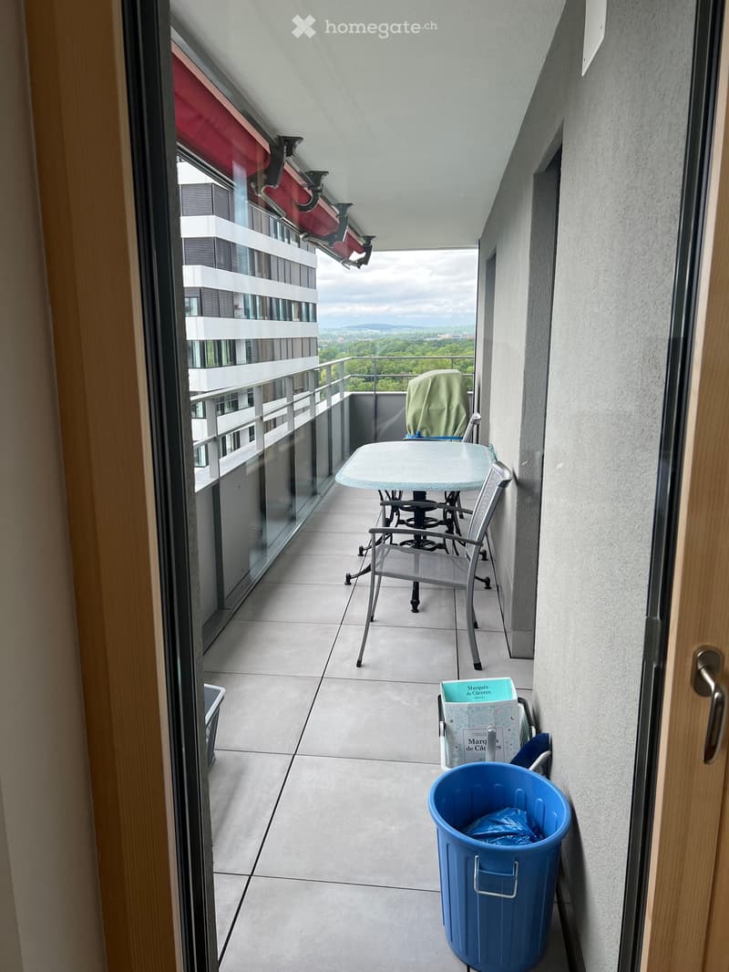 Balcony with a table, two chairs, a covered chair, a blue trash bin, and a view of the city and greenery.