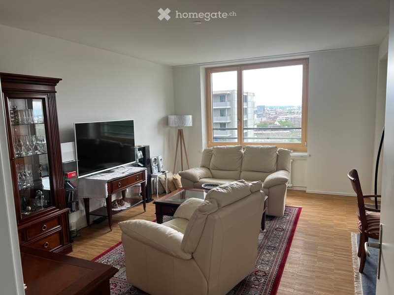 Modern living room with large windows offering a city view, featuring a beige sofa, armchair, coffee table, television, wooden cabinet, and floor lamp.