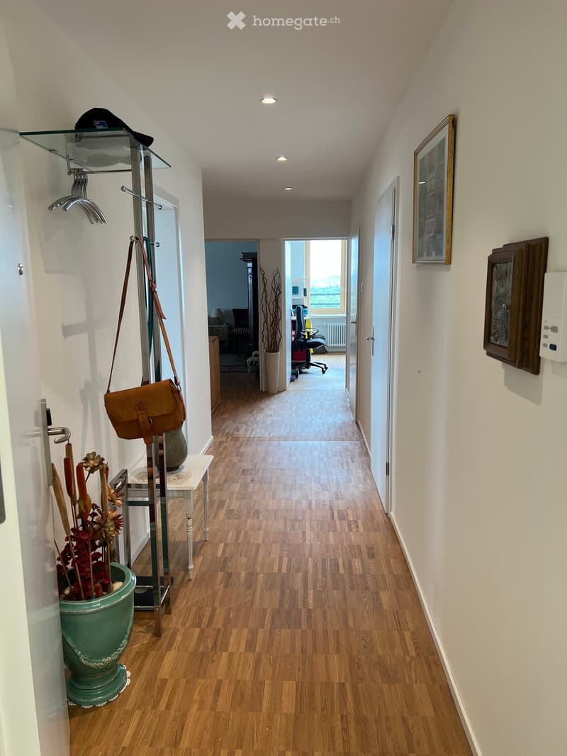 A well-lit hallway with wooden flooring, white walls, and recessed ceiling lights. Features include a coat rack with a bag, a small table with a vase, and framed artwork on the walls. The hallway leads to an open area with a view of a room containing offi