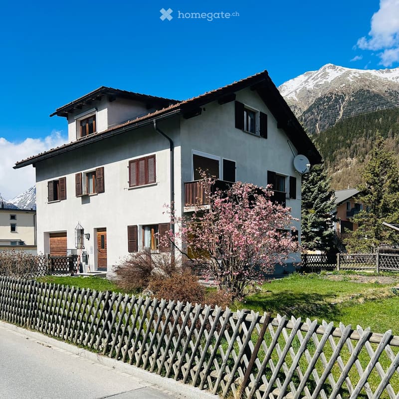 Two-story white house with brown shutters, wooden door, balcony, and surrounding garden with blooming trees and mountains in the background.