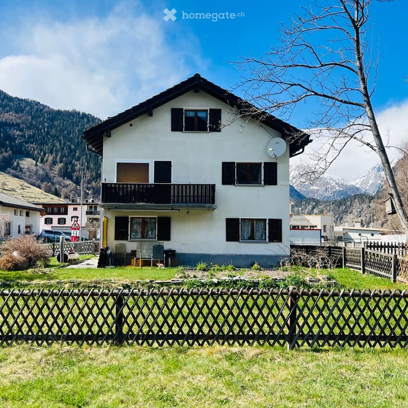 Two-story white house with black shutters and a balcony, surrounded by a wooden fence and green lawn, with mountain views in the background.