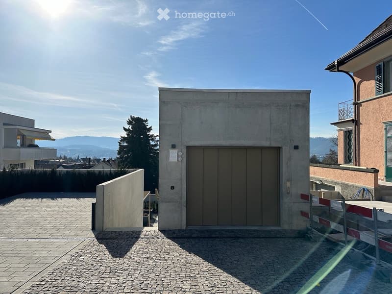 Modern concrete building with a large garage door, surrounded by a paved area with a view of the city and mountains in the background.