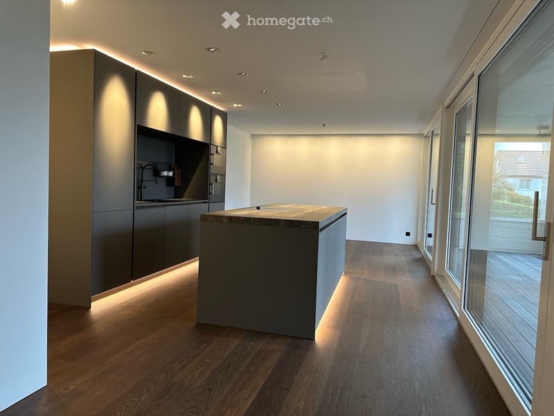 Modern kitchen with dark grey cabinetry, a central island, and wooden flooring. The space features built-in lighting and large windows offering a view of the outdoors.
