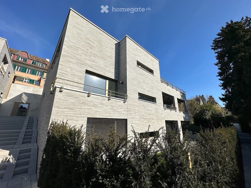 Modern, multi-story building with a sleek, angular design and light-colored facade. Features include large windows, a raised ground floor, and a balcony on the upper level.