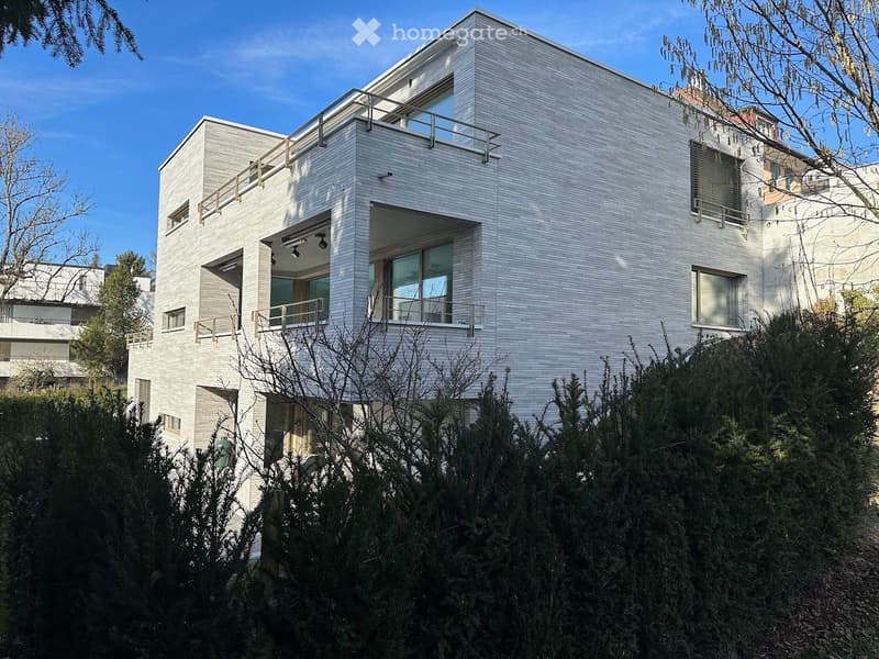 Modern, two-story house with white brick exterior, large windows, and balconies. The house features a sleek, contemporary design with clean lines and is surrounded by greenery.