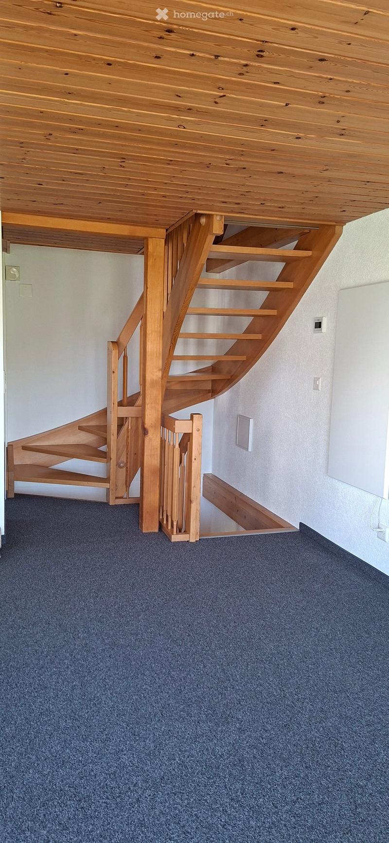Spacious hallway with a wooden staircase featuring open wooden railings and steps, carpeted floor, and white walls.