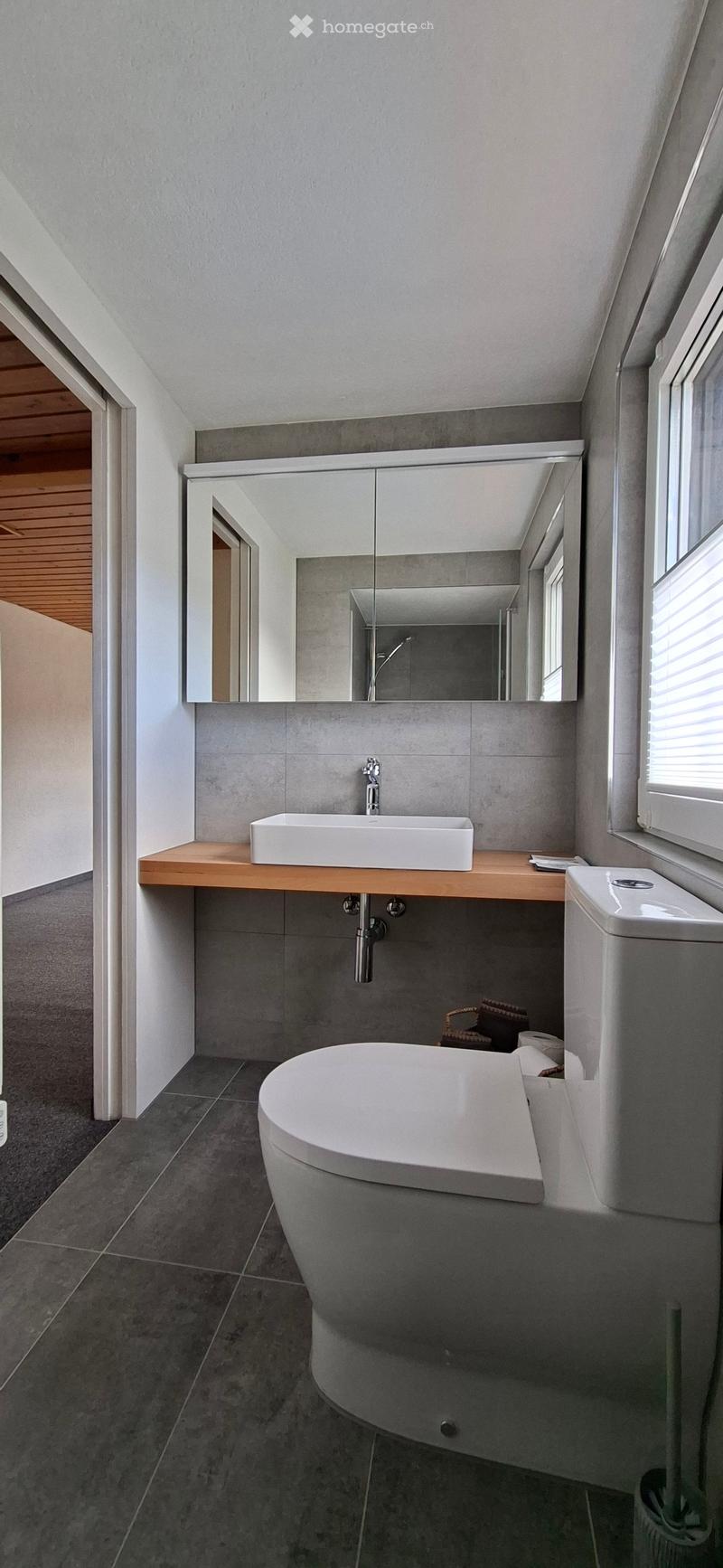Modern bathroom with a white sink, mirror, toilet, and bathtub, featuring grey tiles and a wooden countertop.