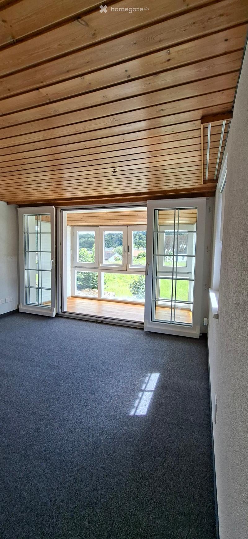 Empty room with grey carpet, wooden slanted ceiling, and large sliding glass doors leading to a wooden deck with a view of greenery.