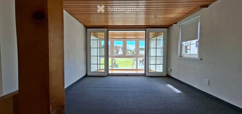 Empty room with grey carpet, light wooden ceiling, large glass doors leading to a patio, and a window with blinds on the right side.