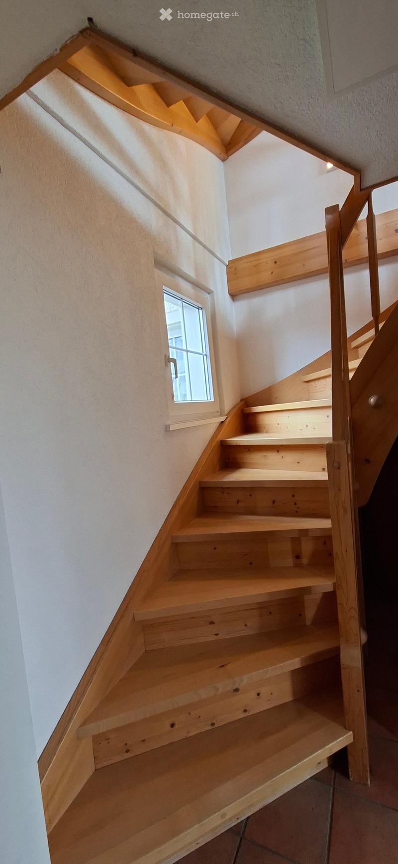 Wooden staircase with light-colored steps and railings, white walls, small window on one side, and a glimpse of another staircase in the background.