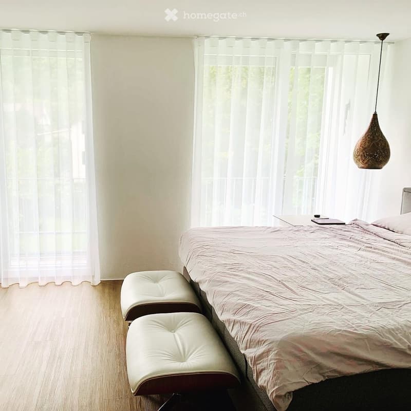 A modern bedroom with a large bed, two white leather stools, a small table with a tablet, and a hanging lamp.