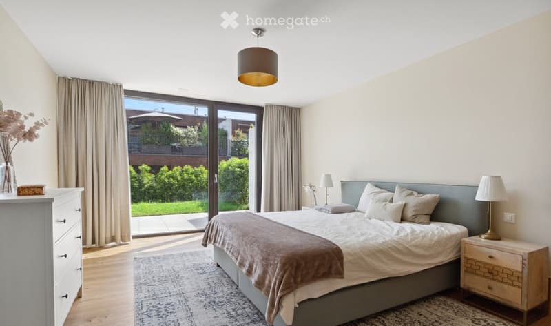Modern bedroom with a large bed, nightstands, lamps, dresser, and a view of a garden through large windows.