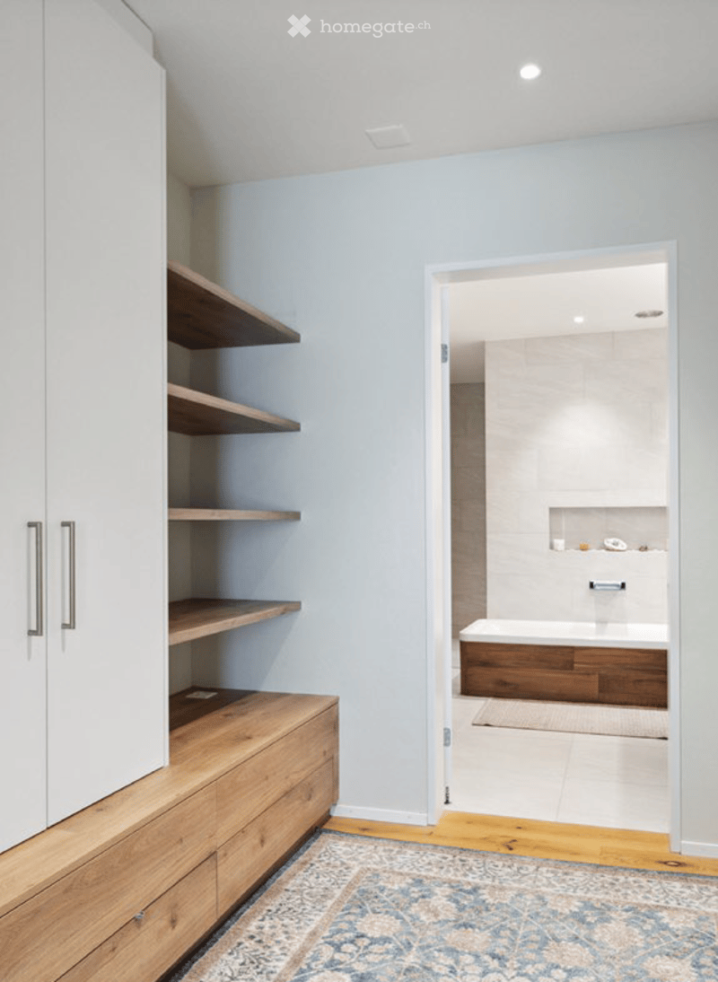 Modern bathroom with a wooden bathtub, built-in wooden shelves, white cabinet, and a patterned rug on the floor.