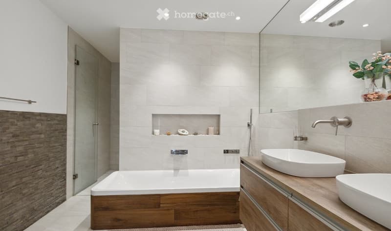 Modern bathroom with a white bathtub, two sinks, a large mirror, and a wooden cabinet. The walls are tiled in light grey, and there is a glass shower enclosure on the left side.
