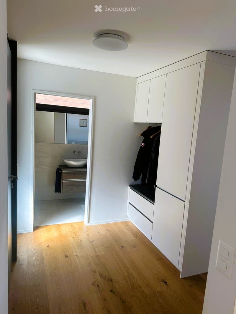 A modern hallway with light wooden flooring, white walls, built-in white storage units, and a small adjacent bathroom visible through an open doorway.