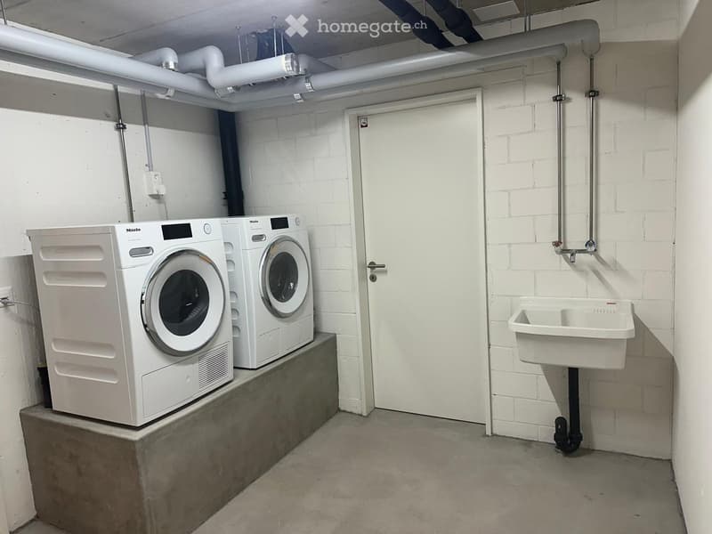 Utility room with a washing machine, dishwasher, and a sink, white brick walls, and a concrete floor.
