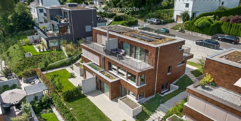 Aerial view of a modern, multi-story residential building with a green roof, balconies, and surrounding landscaped gardens. The building features contemporary architecture with large windows and a mix of wood and concrete materials.