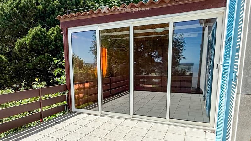 Large sliding glass doors open to a tiled balcony area with a wooden railing and lush greenery in the background.