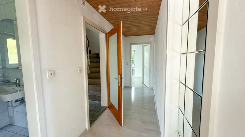 Bright hallway with wooden ceiling, white walls, and light-colored flooring. Features an open wooden door leading to a staircase and a partially visible bathroom on the left side.