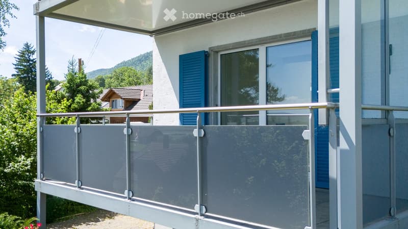 Balcony with glass railing, overlooking a scenic view of trees and a house in the background.