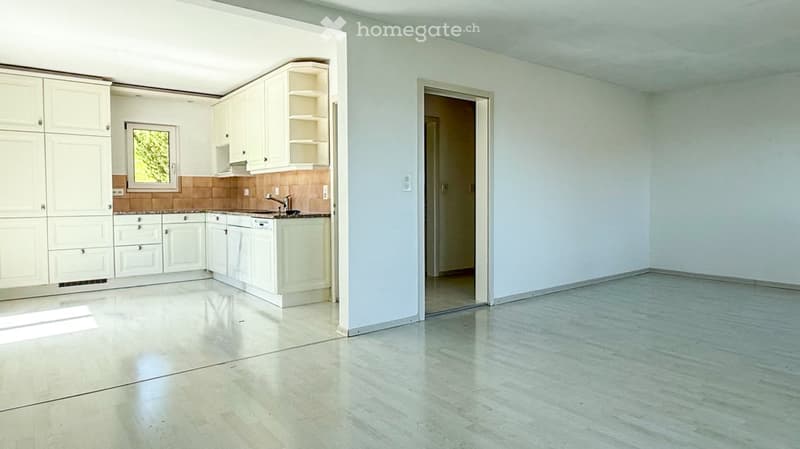 Empty room with white walls, light-colored tiled floor, and an open doorway leading to another room. The adjacent kitchen area features white cabinets, a countertop, and a sink.