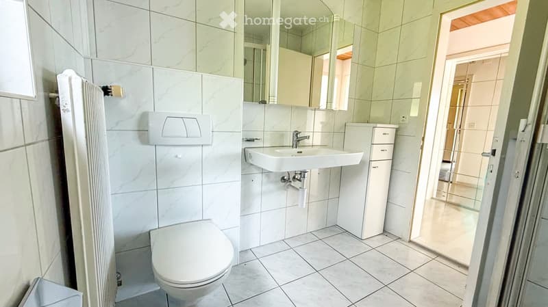 Modern bathroom with white tiles, a toilet, a sink with a mirror, and a small cabinet. The floor is tiled, and the walls are covered in marble-effect tiles.