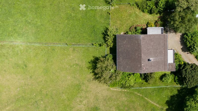 Aerial view of a single-story house with a brown shingled roof, surrounded by a grassy lawn and trees.