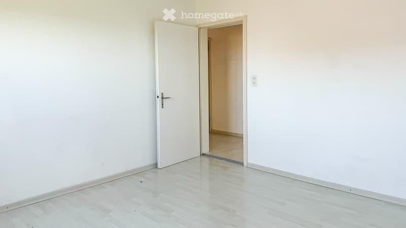 Empty room with white walls, light wooden flooring, and an open door leading to another room.