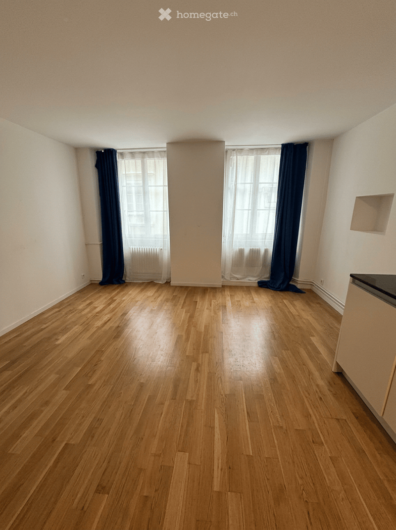 Empty room with wooden flooring, white walls, large windows with dark curtains, and a small built-in shelf on one wall.