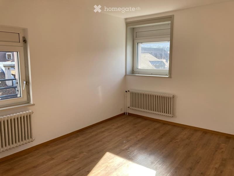Empty room with wooden flooring, white walls, two windows, and radiators on both sides.