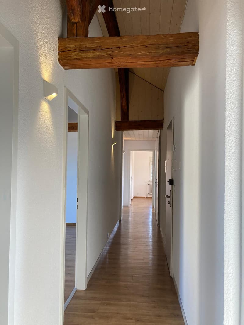 A long, narrow hallway with wooden flooring, white walls, and exposed wooden beams on the ceiling. The hallway features several doors and a wall-mounted light fixture on the left side.