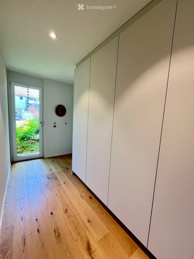 Modern hallway with light wooden flooring, white walls, built-in white cabinets, and a sliding glass door leading to a garden area.