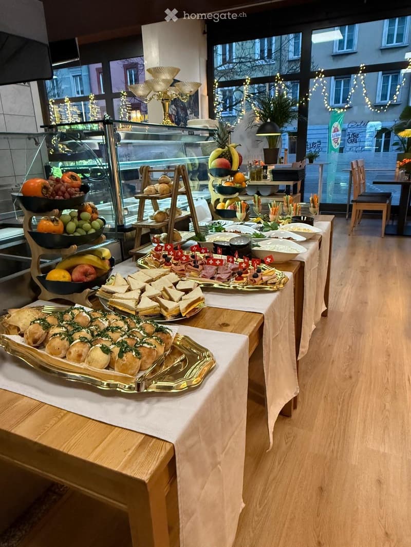 A buffet setup with various food items including sandwiches, fruits, and cakes arranged on tables covered with white tablecloths. The room has large windows with a view of the outside and is decorated with string lights.