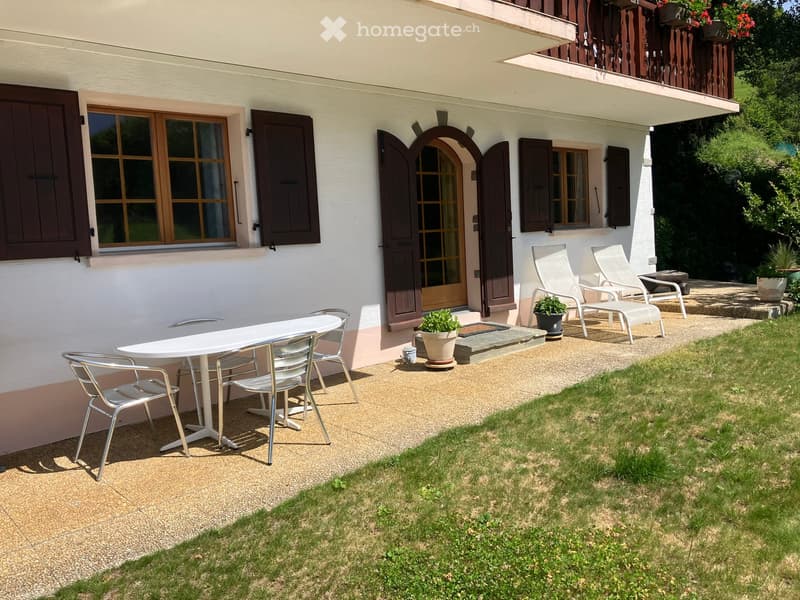 Outdoor patio area with a white table and multiple chairs, a small potted plant, and a grassy lawn. The patio is adjacent to a house with white walls, wooden shutters, and a wooden deck with additional seating.