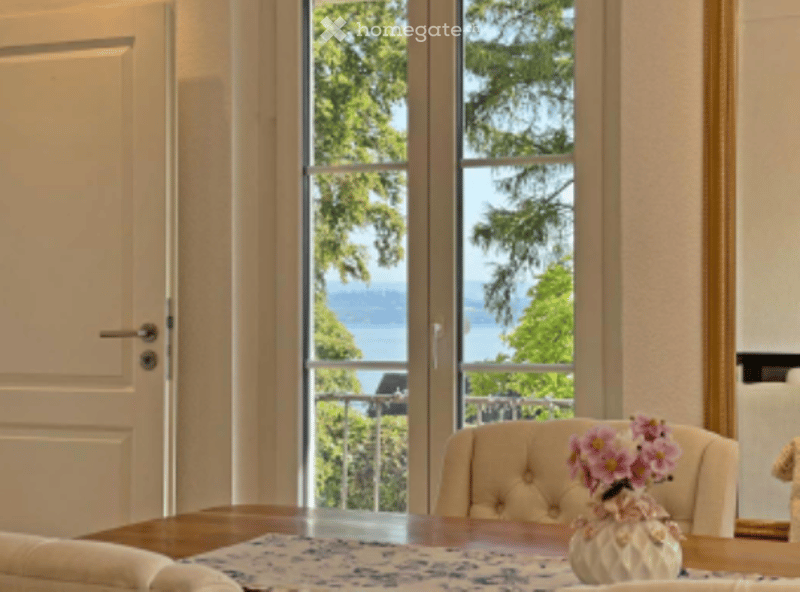 Dining area with a wooden table, a white vase with pink flowers, and a view of the water through large windows.