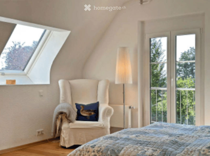 Cozy attic bedroom with slanted ceilings, large window, and skylight. Features a bed with patterned bedding, a white armchair with a blue pillow, and a tall floor lamp.