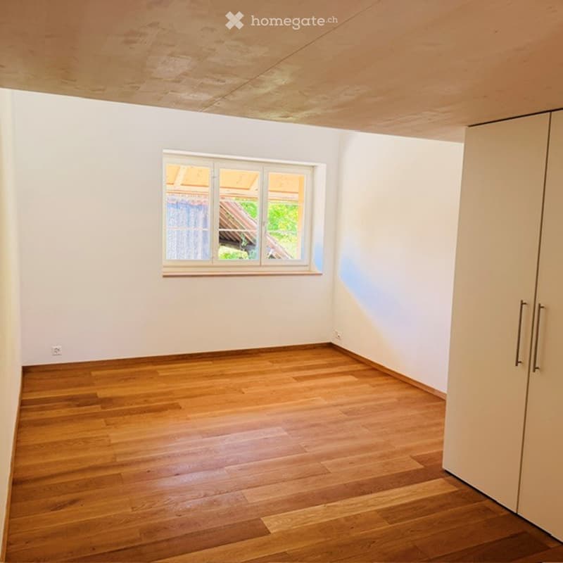 Empty room with wooden flooring, white walls, and a single window with a view of a roof and greenery outside.
