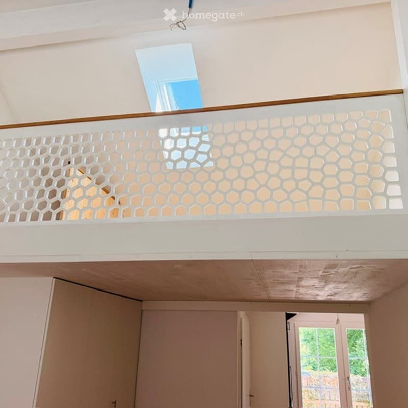 A modern hallway with a white ceiling, a decorative balcony railing featuring a geometric pattern, and a view of a room below.