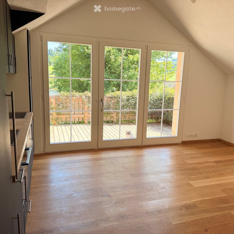 Empty room with wooden flooring, large sliding glass doors leading to a wooden deck, and a view of greenery outside.