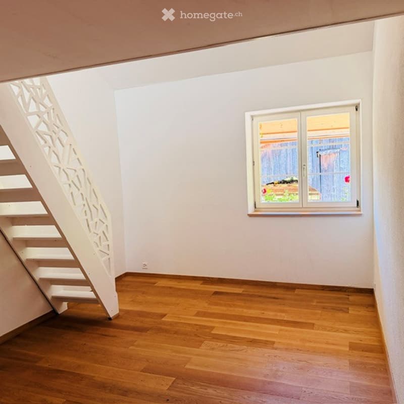 Empty room with wooden flooring, white walls, and a staircase leading upwards. A window with white frames is visible on one wall, showing a view of the outside area.