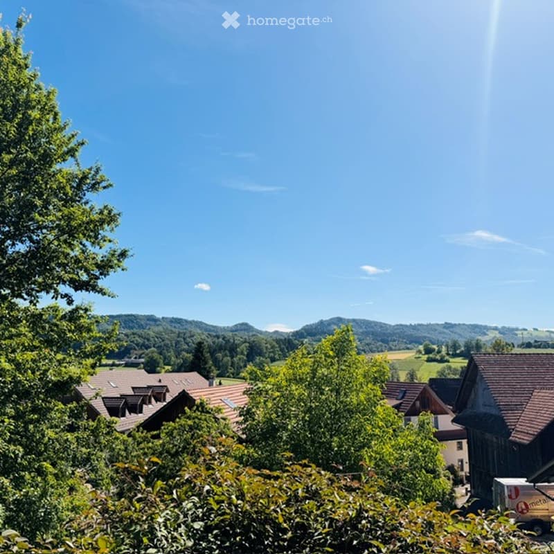 A scenic view of a rural village with traditional houses, surrounded by lush green trees and rolling hills under a clear blue sky.