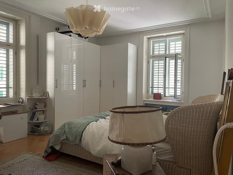 A well-lit bedroom with a large bed, a wicker chair, a wardrobe, a table lamp, a fan, a bookshelf, and a laptop on a desk.