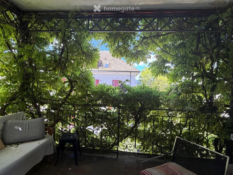 A covered balcony with a metal railing, lush greenery, a sofa, a bench, and a small stool.