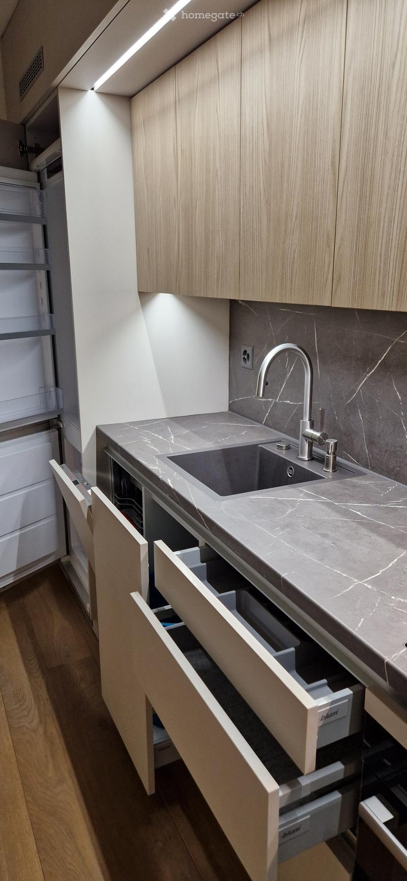 Modern kitchen with a marble countertop, stainless steel sink, and light wood cabinets. Drawers are open, revealing storage compartments.