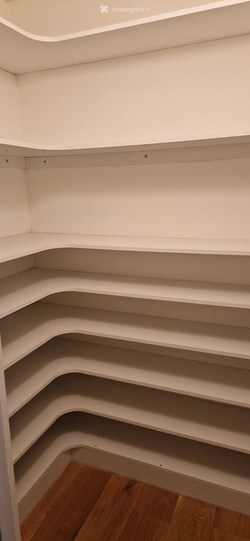 Empty corner closet with white built-in shelves and wooden floor.