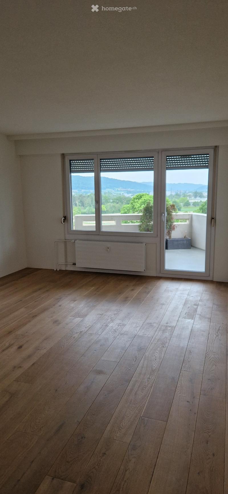 Empty room with wooden flooring, large windows, and a sliding door leading to a balcony with a scenic mountain view.