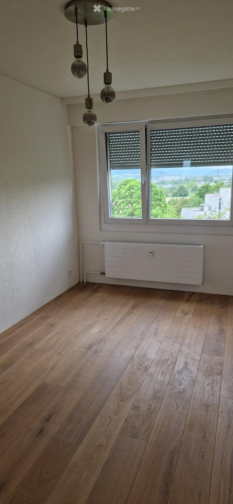 Empty room with wooden flooring, white walls, a radiator, and a window with blinds offering a view of greenery and buildings.